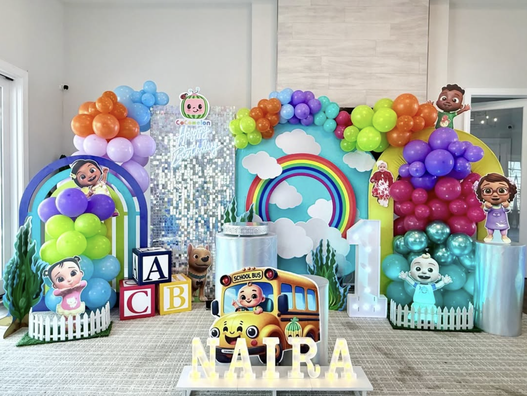 Colorful first birthday celebration with balloons, themed panels, and illuminated name letters